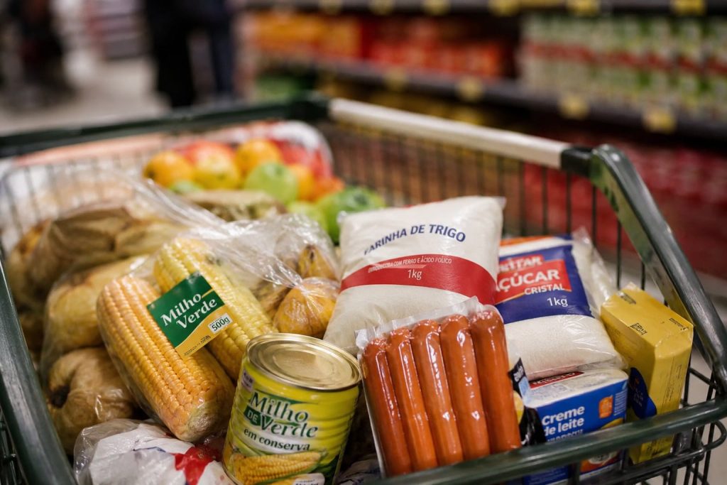 Imagem mostra carrinho de supermercado com comidas típicas de festa junina, como milho, salsicha e açúcar