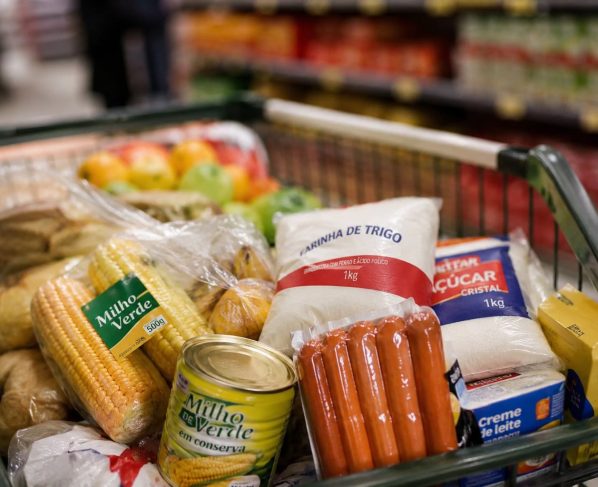 Imagem mostra carrinho de supermercado com comidas típicas de festa junina, como milho, salsicha e açúcar